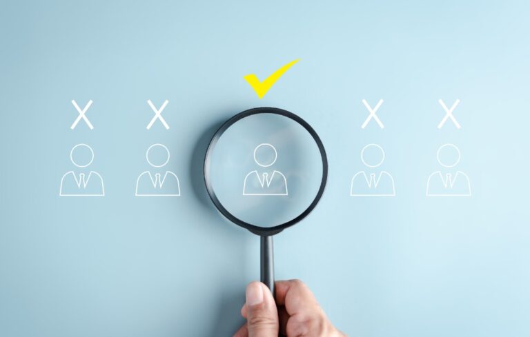 Hand holding magnifying glass over a line of outlines of people with a checkmark over one
