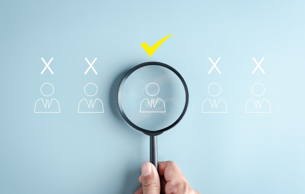 Hand holding magnifying glass over a line of outlines of people with a checkmark over one