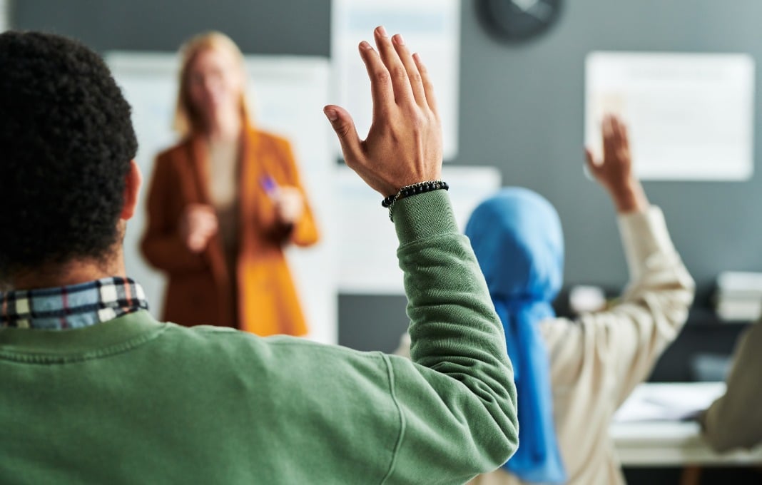 Part of back of young male student in green pullover raising hand at lesson to ask question to teacher or give his answer